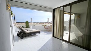 A balcony with white tiled flooring, two lounge chairs, a small table with chairs, and a view of the sea and buildings. - Albufeira Blue Wave Retreat 65 (Albufeira)