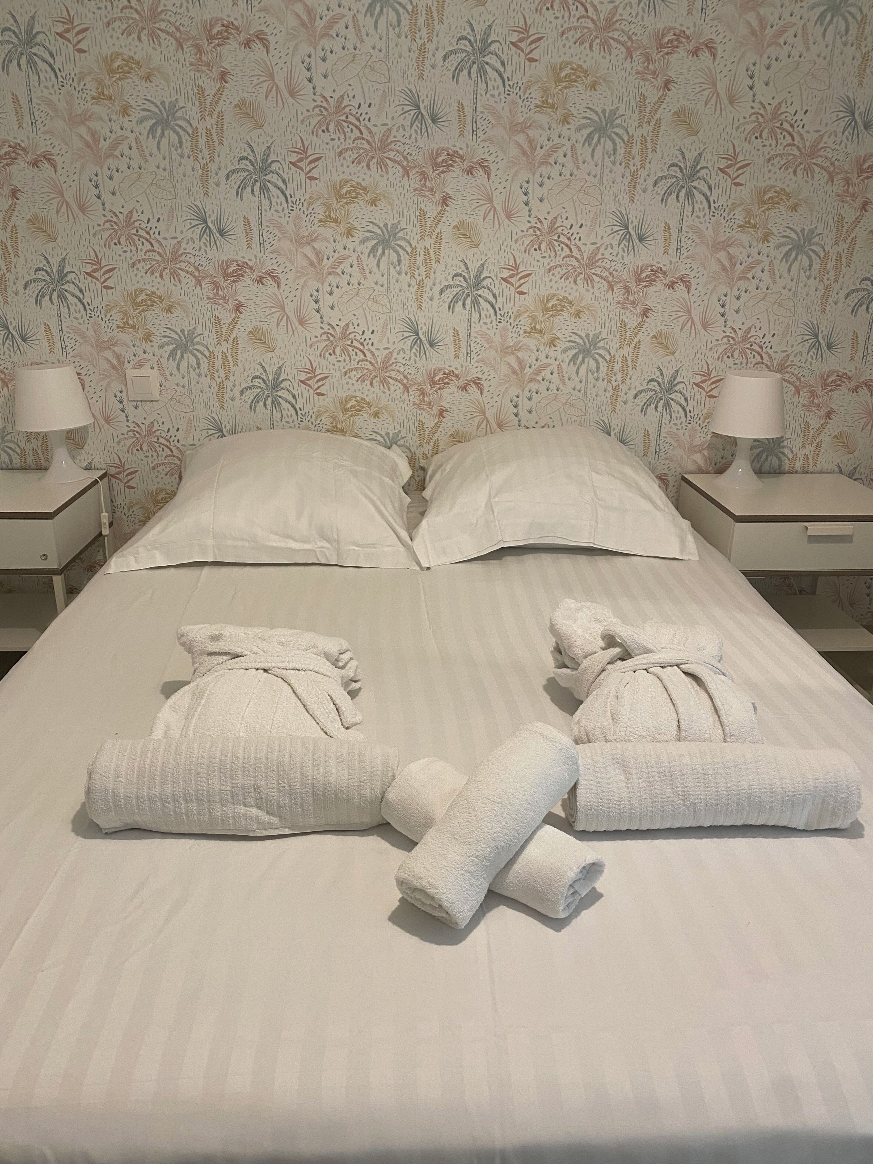 A neatly made bed with white linens and towels, two bedside lamps, and a floral-patterned wallpaper.
