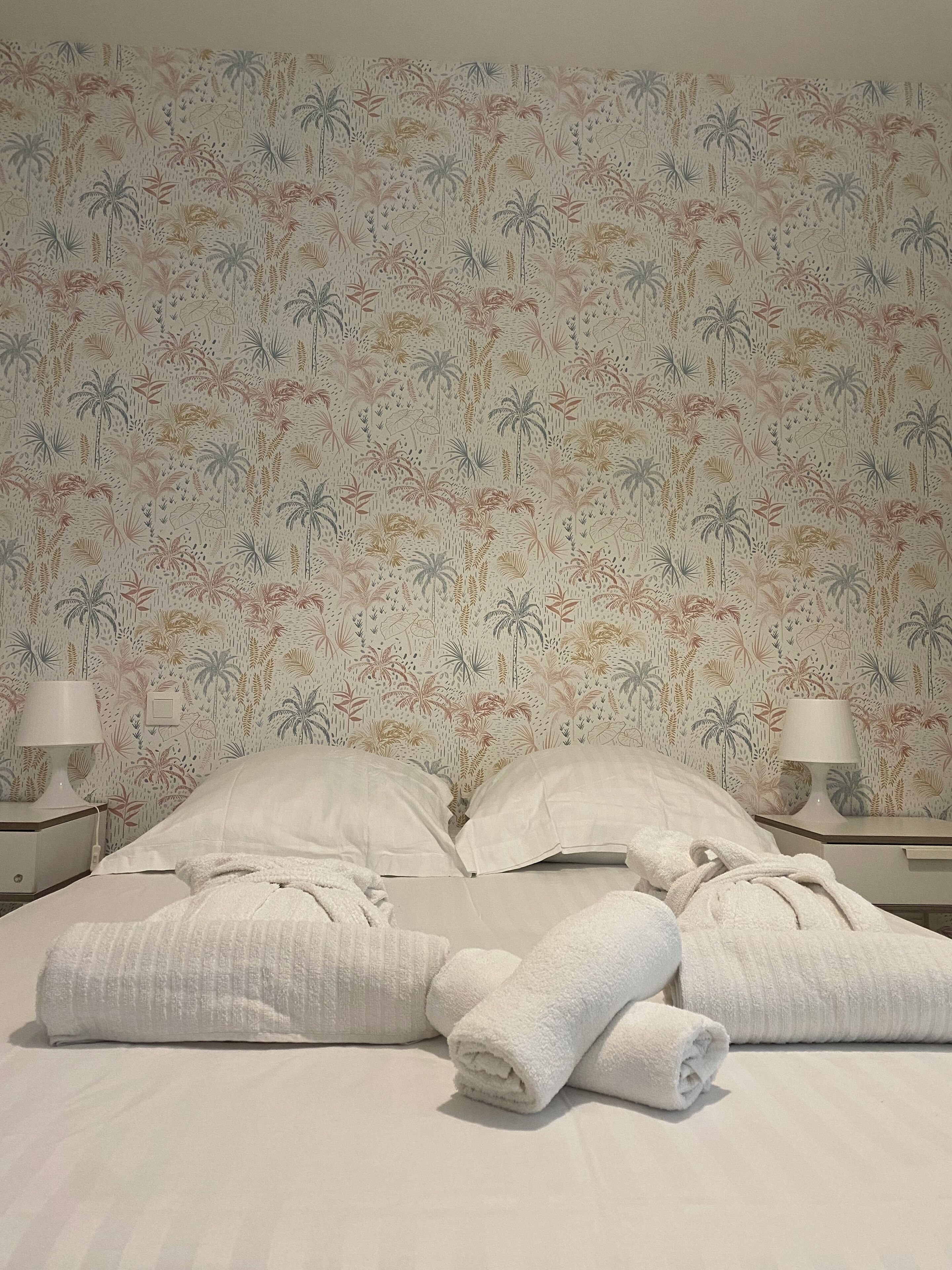Una cama bien hecha con sábanas blancas y dos toallas enrolladas, una mesita de noche con lámpara, y una pared con estampado de plantas tropicales.