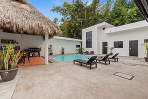 Un espace piscine extérieur moderne, doté de chaises longues, d’un toit de chaume et de plantes en pot.