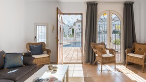Una sala de estar con un sofá, dos sillas de mimbre, una mesa baja y vistas a una piscina a través de una puerta abierta.