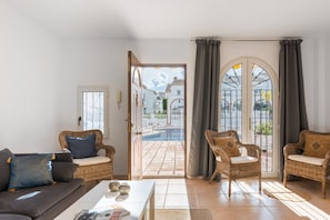 A living room with a sofa, two wicker chairs, a coffee table, and a view of a swimming pool through an open door.