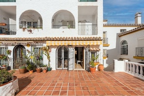 A white building with terracotta tiled ground, arched windows, and potted plants.