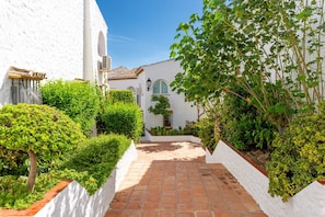 A courtyard with a white building, a tiled pathway, and lush greenery.