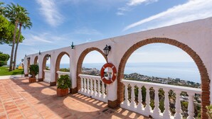 Una terraza con barandilla blanca, arcos de ladrillo, plantas en macetas y vista al mar.