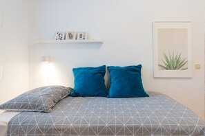 A bedroom with a bed, pillows, a wall-mounted shelf with decorative items, and a framed plant.