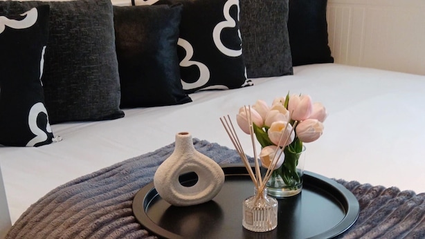 A neatly made bed with a brown textured blanket, a tray with a vase of flowers and reed diffusers, and black and white patterned pillows.