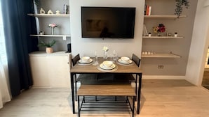 A modern living room with a dining table set for four, a flat-screen TV mounted on the wall, and built-in shelves with decorative items and plants.
