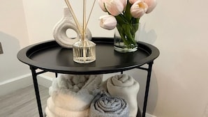 A black metal table with a glass vase containing pink tulips and reed sticks on top. Below, there are rolled blankets in a grey basket.
