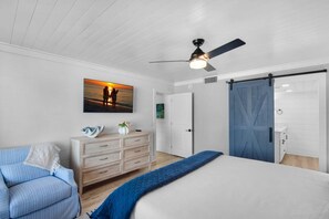 Une chambre à coucher avec un lit, une commode, un canapé bleu, un ventilateur de plafond et un tableau représentant un coucher de soleil.