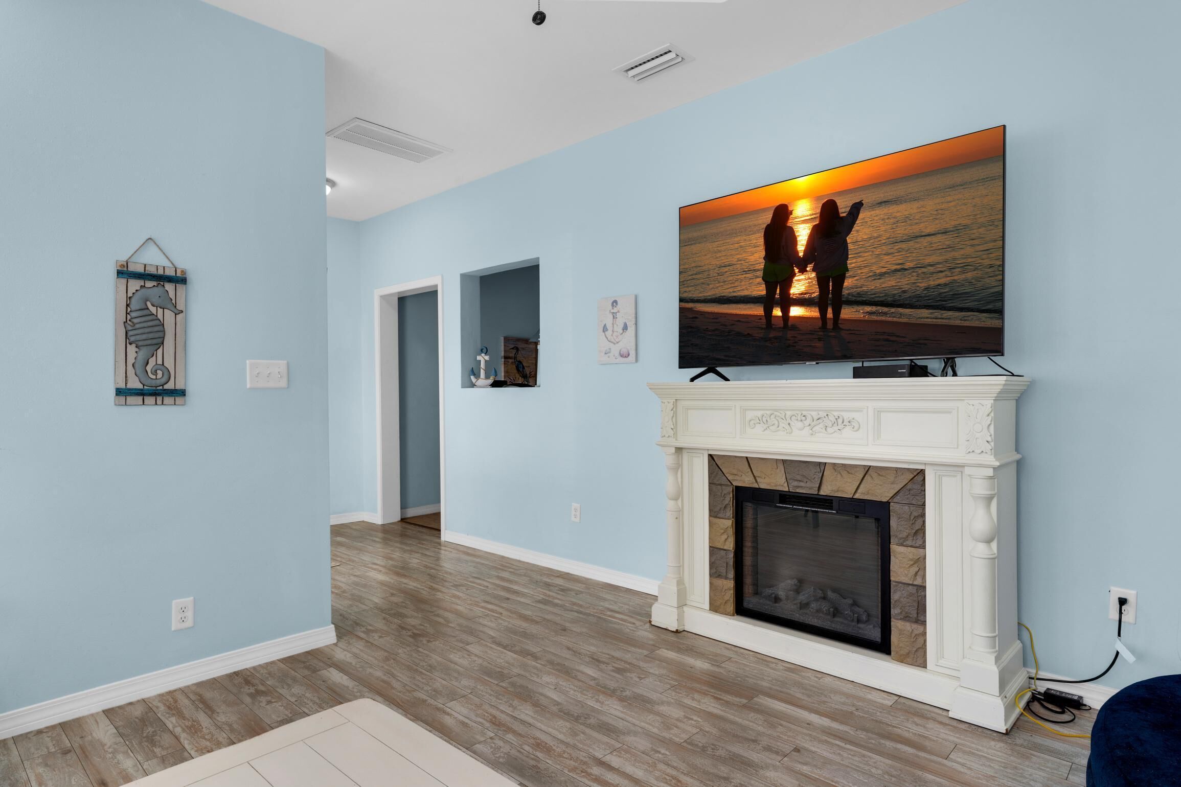 A living room with a fireplace, a large TV displaying a sunset scene, and a hallway leading to another room.