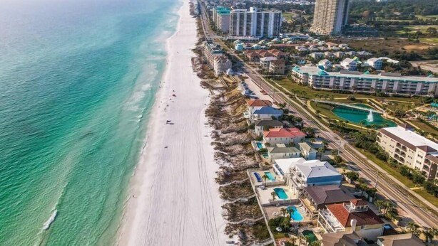 Vista aérea de un complejo turístico frente al mar, con propiedades en la playa, un gran hotel y un conjunto de piscinas.