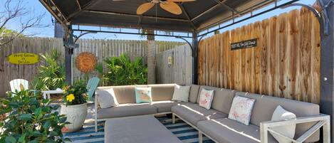 An outdoor seating area with a sectional sofa, a ceiling fan, and a wooden fence.