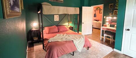 A bedroom with a four-poster bed, a nightstand with a lamp, a desk with a mirror, and a door leading to another room.