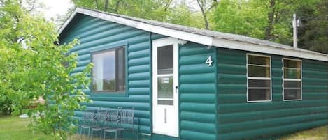 A small, single-story log cabin with a door and windows, numbered 4, surrounded by greenery.