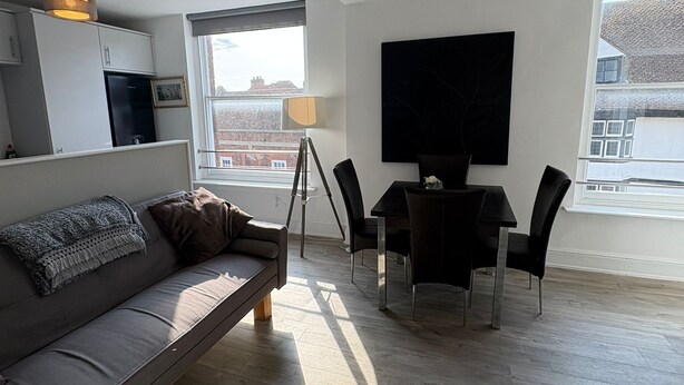 Een moderne woonkamer met een bank, eettafel en stoelen. Er is een televisie aan de muur bevestigd en een raam zorgt voor natuurlijk licht.