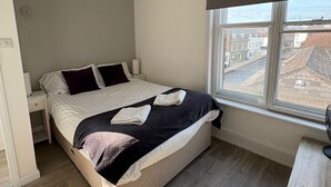 Een moderne slaapkamer met een groot bed, een nachtkastje, een raam met uitzicht op gebouwen en een plafondlamp.