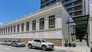 A building with a white facade and large windows, a parking lot with several cars, and a modern building in the background. - Villes de France - Centro de Floripa (Florianopolis)