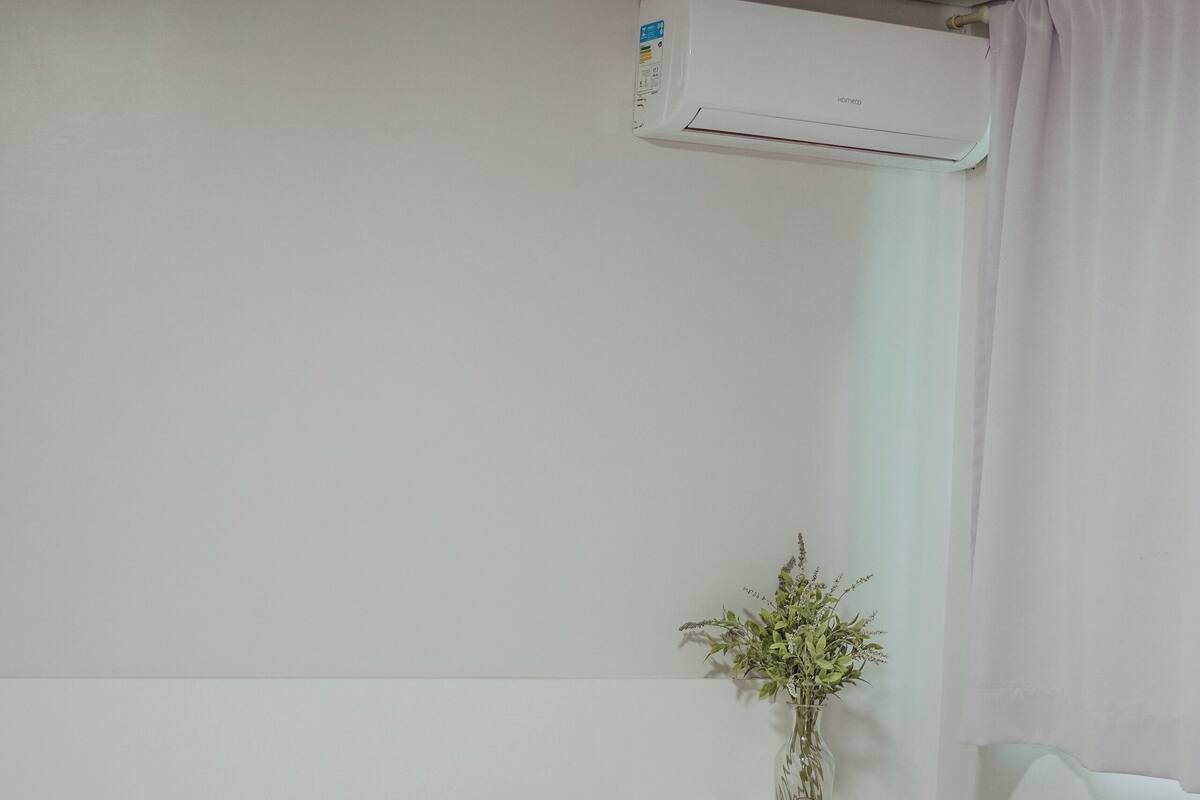 A hotel room with a bed, a nightstand, a vase with flowers, and an air conditioning unit.