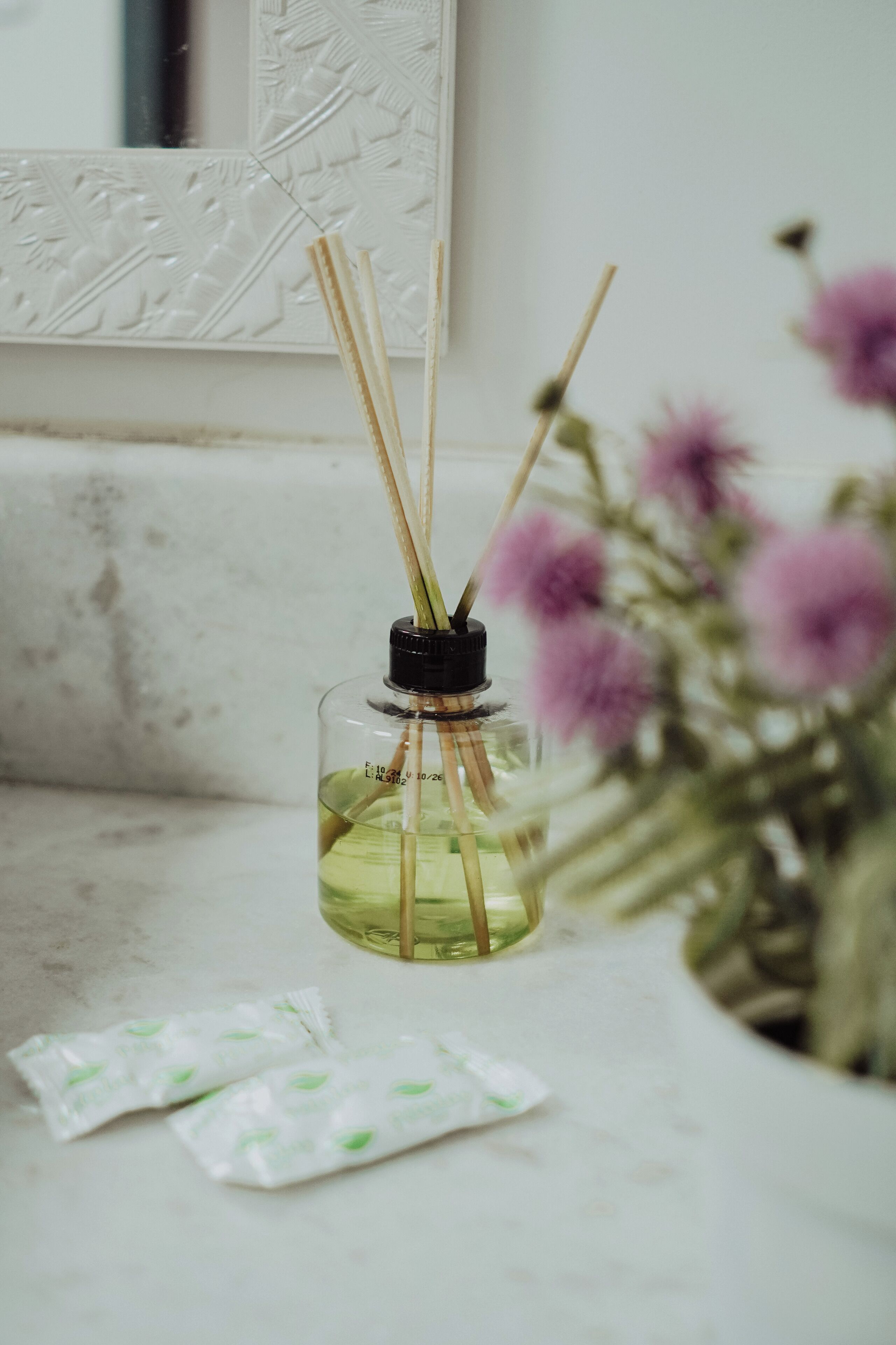 A bottle of reed diffuser with a black cap and reed sticks, placed on a marble surface next to a vase with purple flowers.