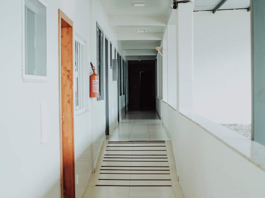 Un long couloir étroit aux murs blancs, avec un extincteur fixé au mur et une porte sur le côté gauche.