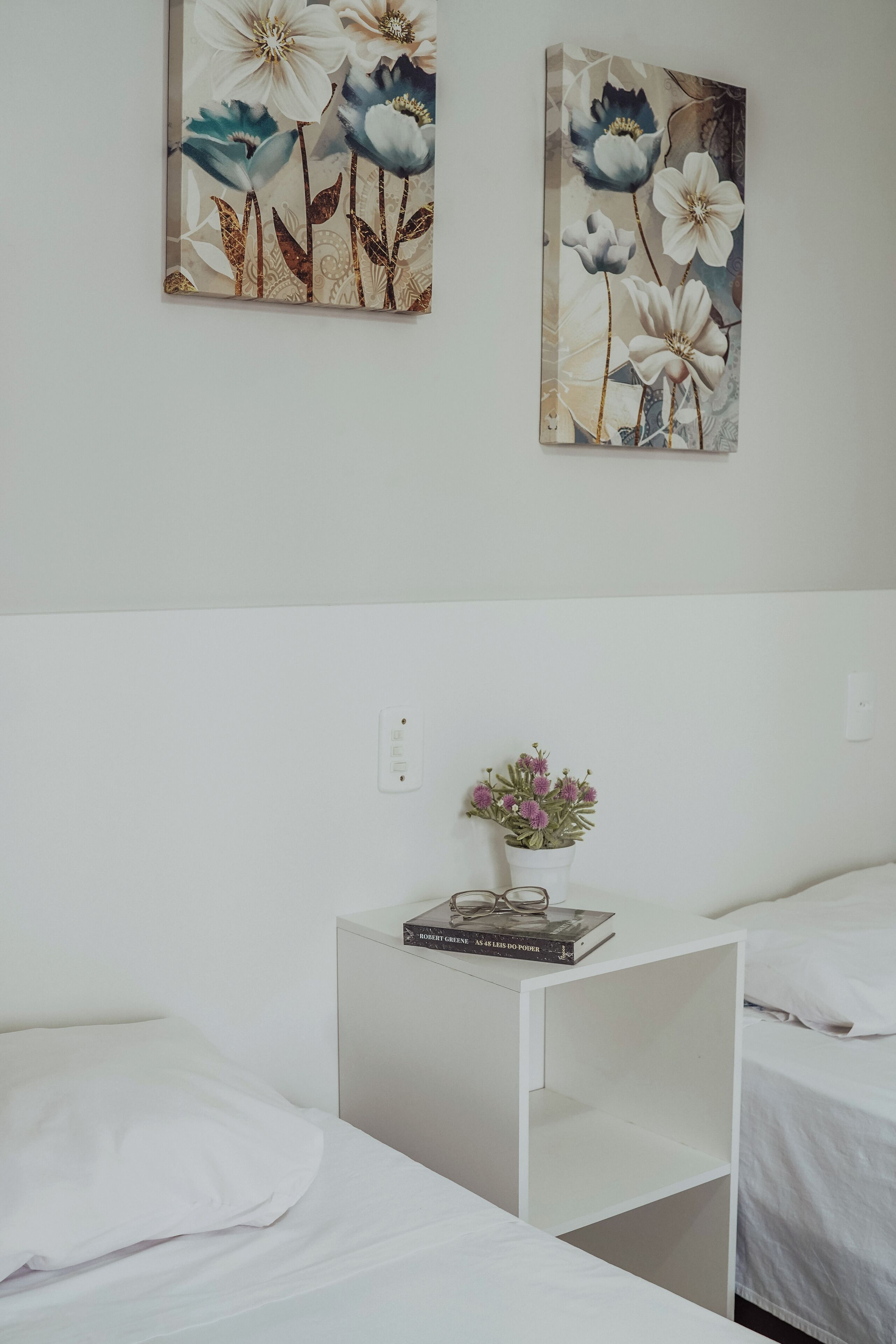 A hotel room with two beds, a white nightstand with a book and a potted plant, and two floral paintings on the wall.