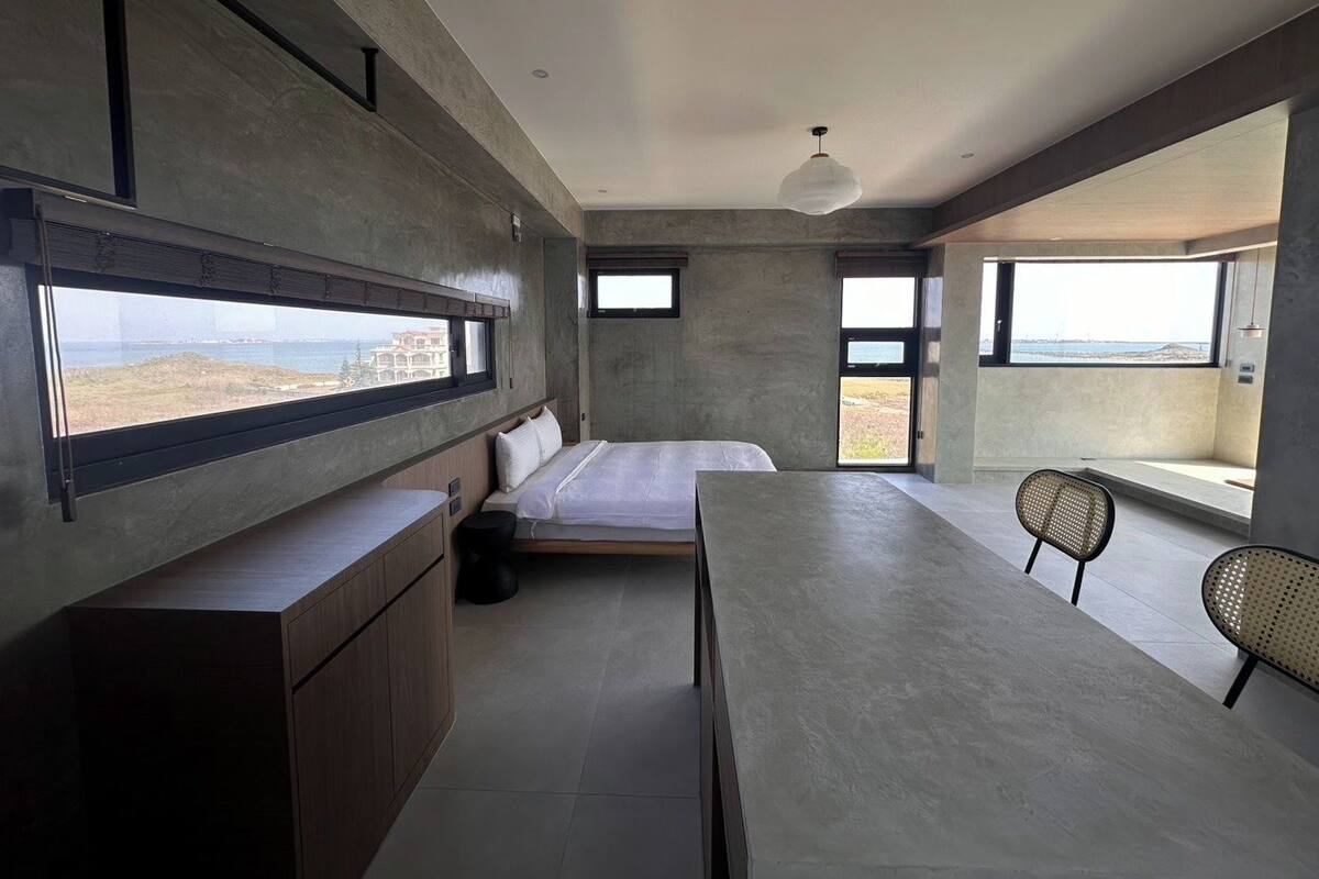 A modern interior with a bed, a dining table, and a seating area with a view of the sea.