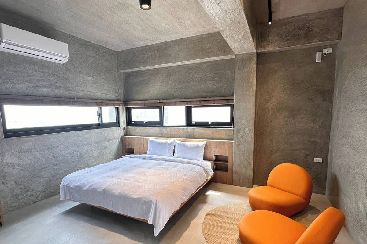 A modern bedroom with a large bed, two windows, an air conditioning unit, and an orange chair.