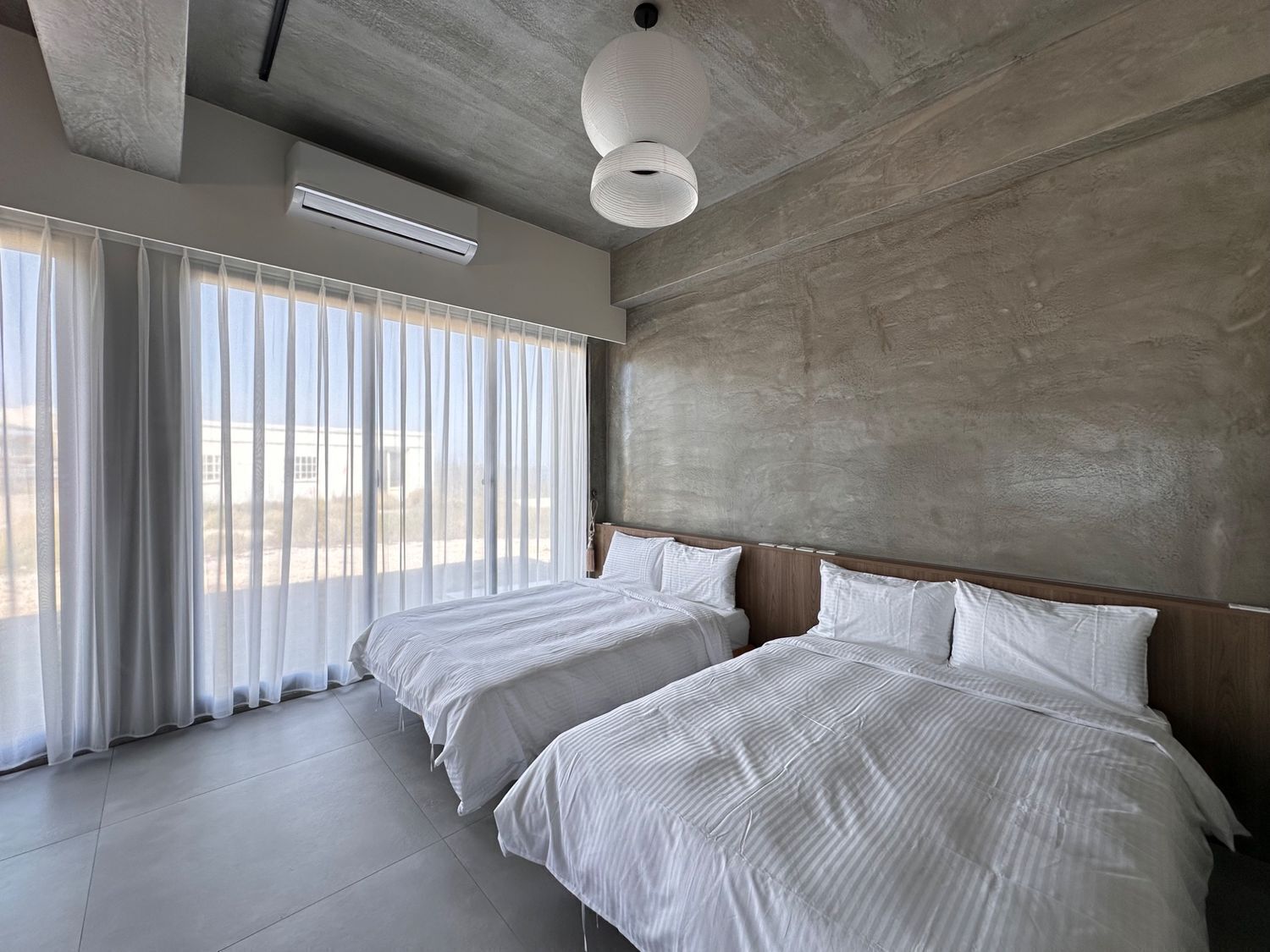 A modern hotel room with two beds, large windows with sheer curtains, a concrete accent wall, and a ceiling-mounted light fixture.
