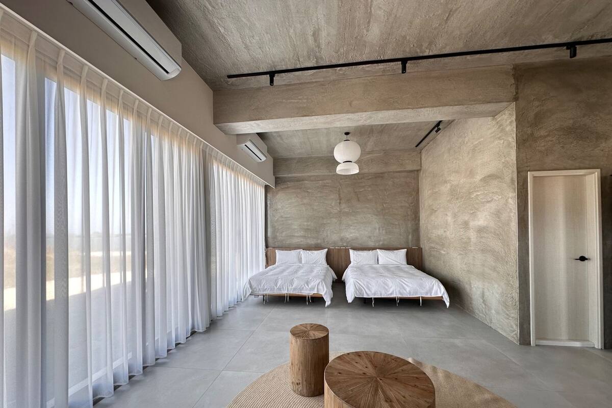 A modern hotel room with two beds, large windows with sheer curtains, a concrete ceiling, and a minimalist design.