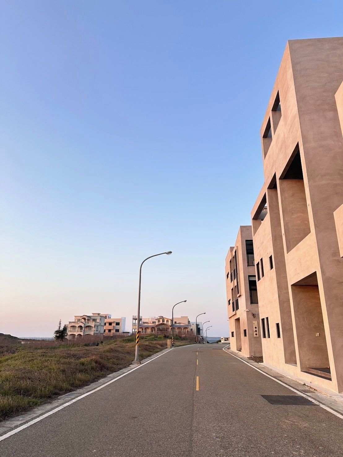 A modern building with a flat facade and multiple balconies, situated beside a road with street lamps.