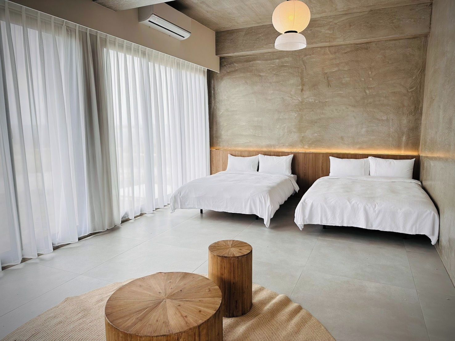 A modern bedroom with two beds, a large window with sheer curtains, a round wooden table, and a textured wall.