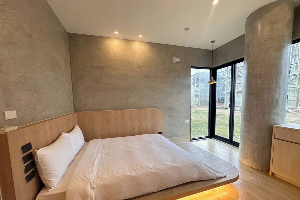 A modern bedroom with a large bed, a wooden headboard, and a view of a construction site through a large window.