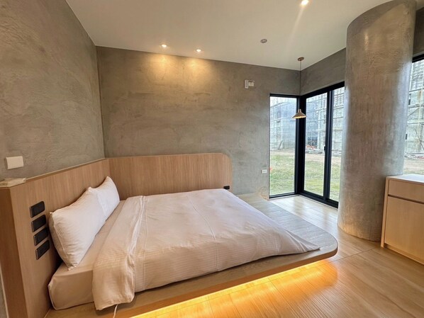 Un dormitorio moderno con una cama grande, cabecera de madera y vista a una obra en construcción a través de un ventanal.