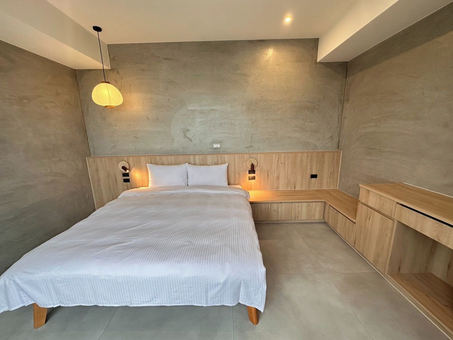 A modern bedroom with a large bed, wooden headboard, bedside tables, and a hanging light fixture.