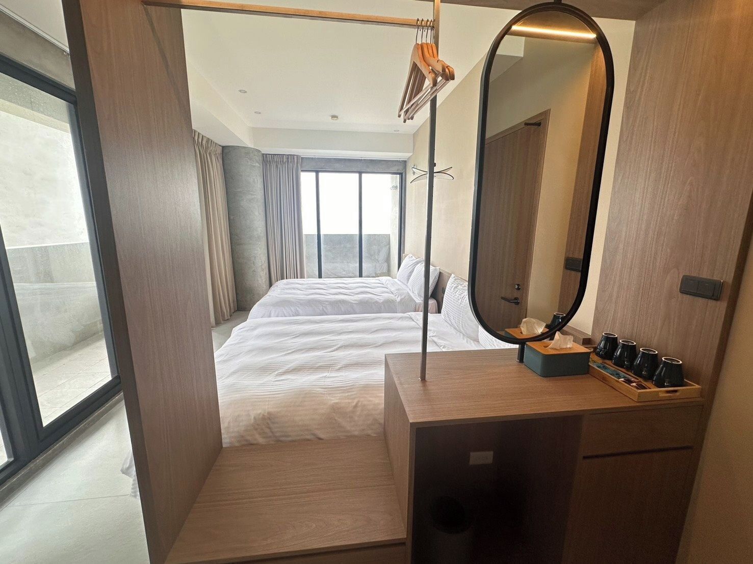 A modern hotel room with a large bed, a wooden vanity with a mirror, and a view of the city through large windows.