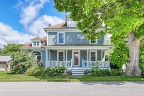 A two-story house with a porch, a front door, and a large tree in front. - The Farmhouse at South Hero (South Hero)