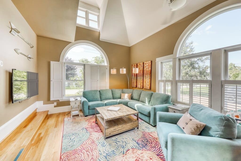 A spacious living room with a large window, a sofa set, a coffee table, and a TV mounted on the wall.