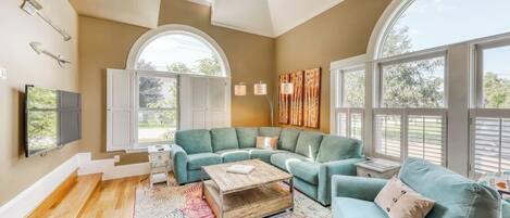 A spacious living room with a large window, a sofa set, a coffee table, and a TV mounted on the wall.