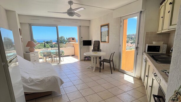 A hotel room with a bed, a desk, a chair, a TV, a ceiling fan, and a kitchen area. - St Clair - Sea view apartment (Le Lavandou)