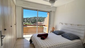 A bedroom with a bed, a ceiling fan, and a view of a residential area through a sliding door. - St Clair - Sea view apartment (Le Lavandou)