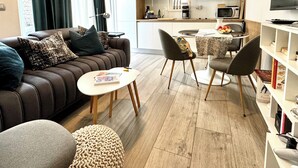 Ein modernes Wohnzimmer mit einer Couch, einem Couchtisch und einem Hocker. Es gibt einen Essbereich mit Tisch und Stühlen. Im Hintergrund ist eine Küche mit Geräten und einem Fenster erkennbar.