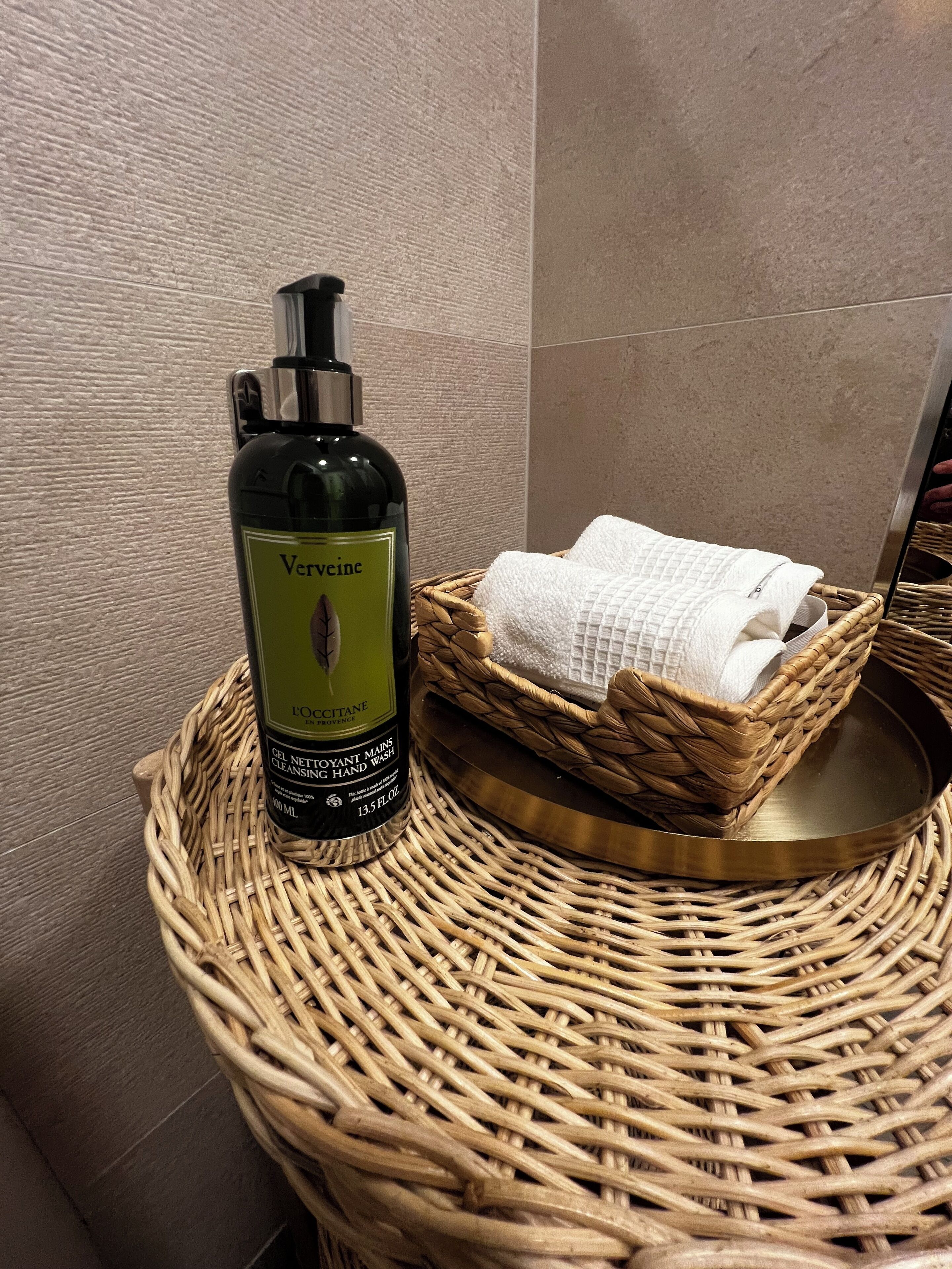 A bottle of L'OCCITANE hand wash and a wicker basket with folded towels on a round wicker table in a bathroom.