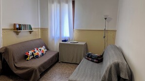 Una habitación pequeña y acogedora con un sofá, una mesa pequeña con un plato y una taza, una lámpara y una estantería llena de libros.