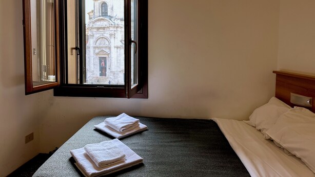 Una habitación con una cama, una ventana que ofrece vista despejada de un edificio y dos toallas dobladas sobre la cama.