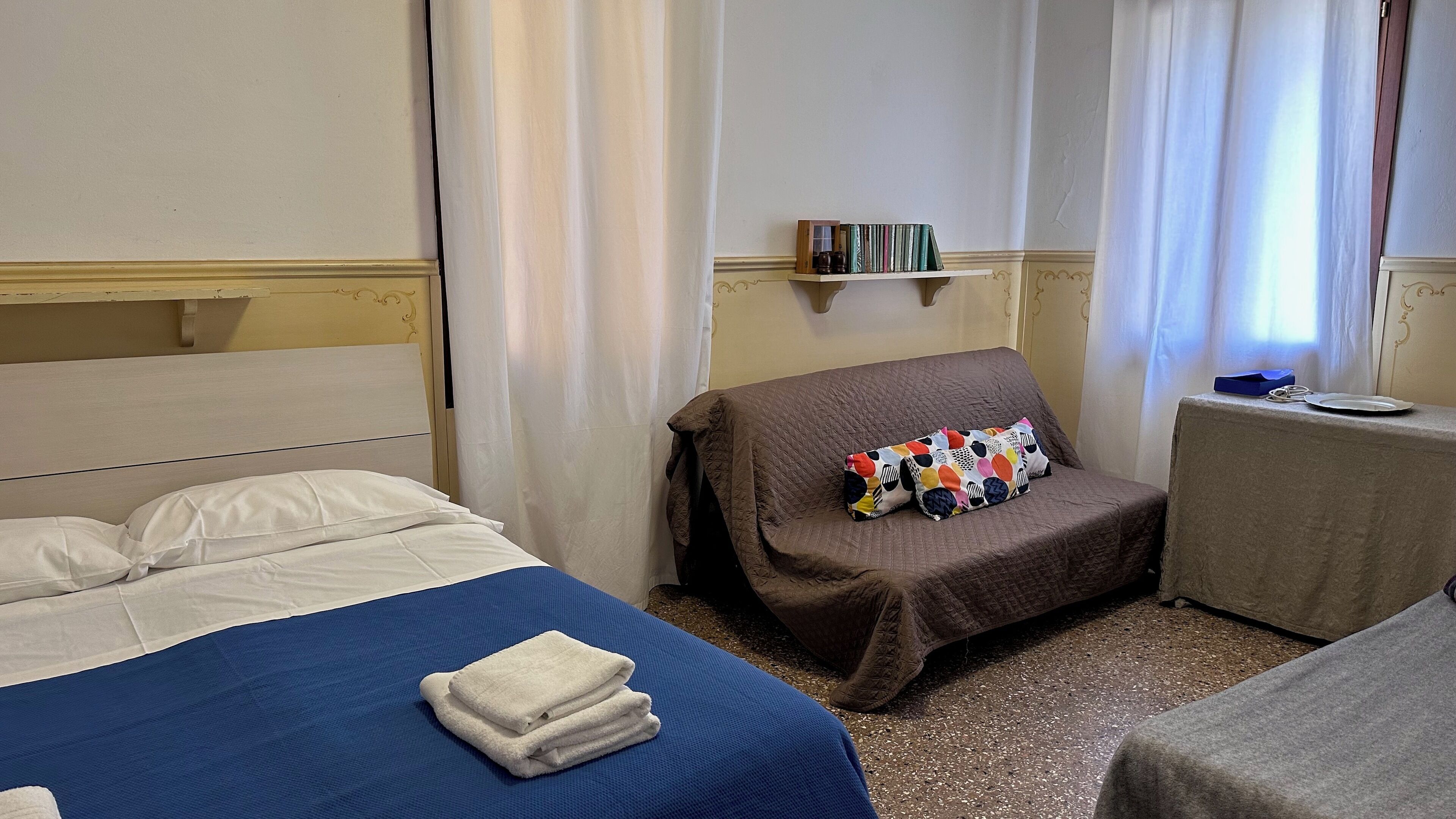 A hotel room with a bed, a small bedside table, a chair, a shelf with books, and a window with curtains.