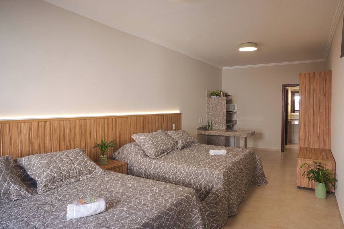 A hotel room with two beds, a wooden headboard, a small bedside table with a plant, a built-in shelf with a microwave, and a wooden wardrobe.