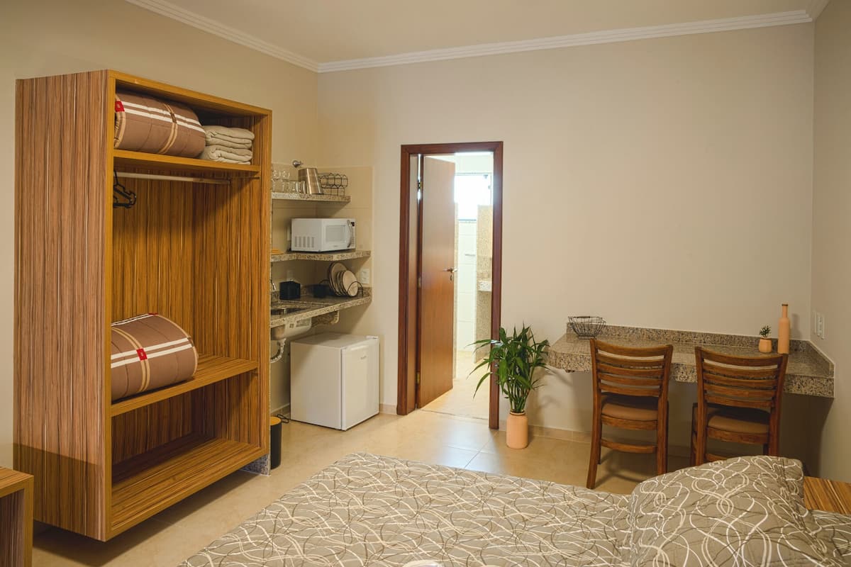 A bedroom with a wooden wardrobe, a bed with patterned bedding, a small kitchenette, and a potted plant.