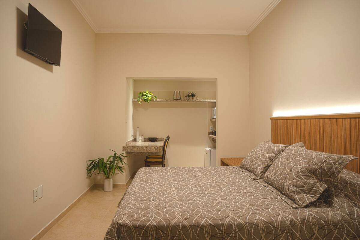 A bedroom with a bed, a bedside table, a nightstand, a TV mounted on the wall, and a small kitchen area with a sink and a plant.