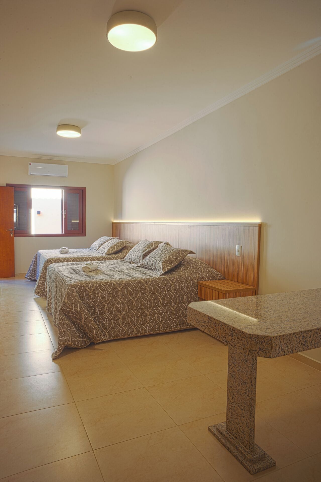 A hotel room with two beds, a wooden headboard, a marble bench, and a ceiling light.